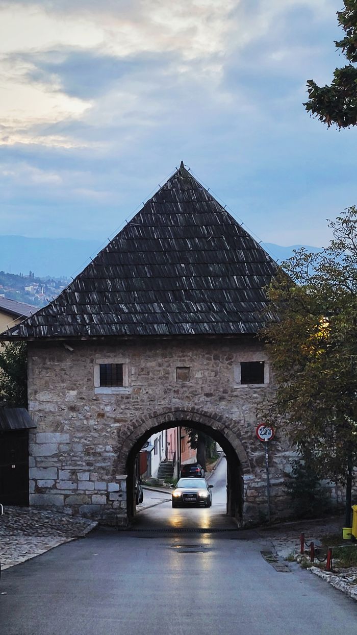 A car drives through an old stone gatehouse, surrounded by trees, under an early evening sky.